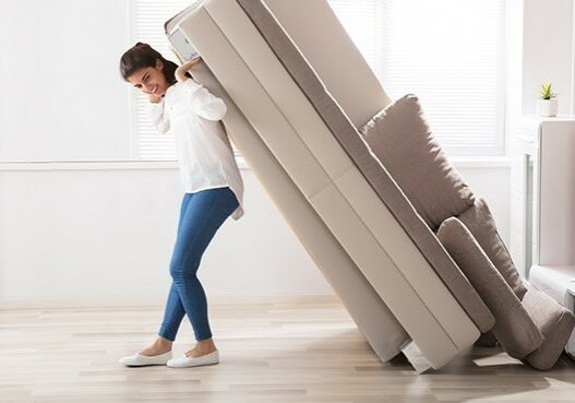 Smiling Young Woman Trying To Move Large Sofa At Home